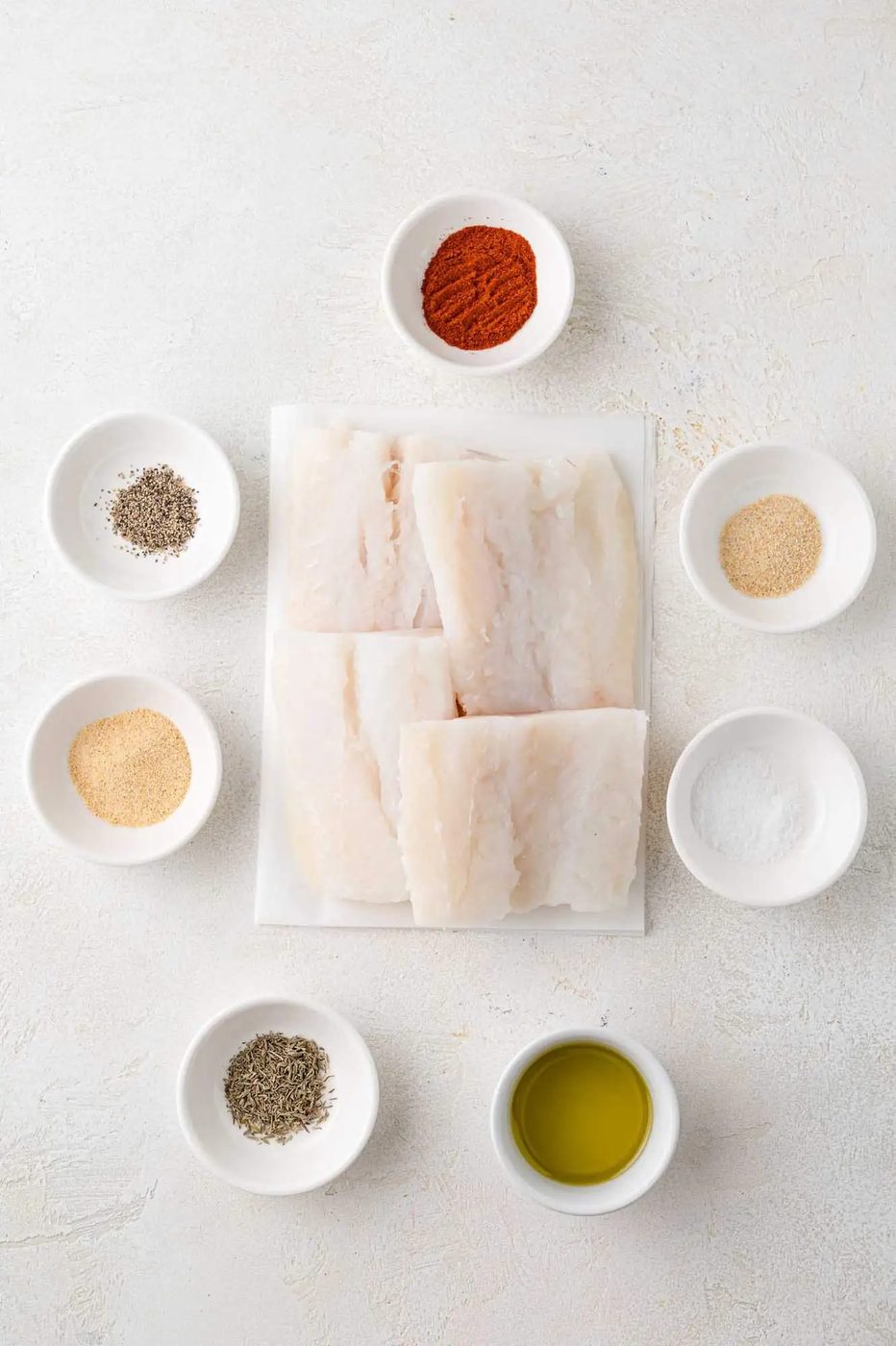 Raw cod fillets on a plate surrounded by small bowls of spices, herbs, and olive oil on a white surface.