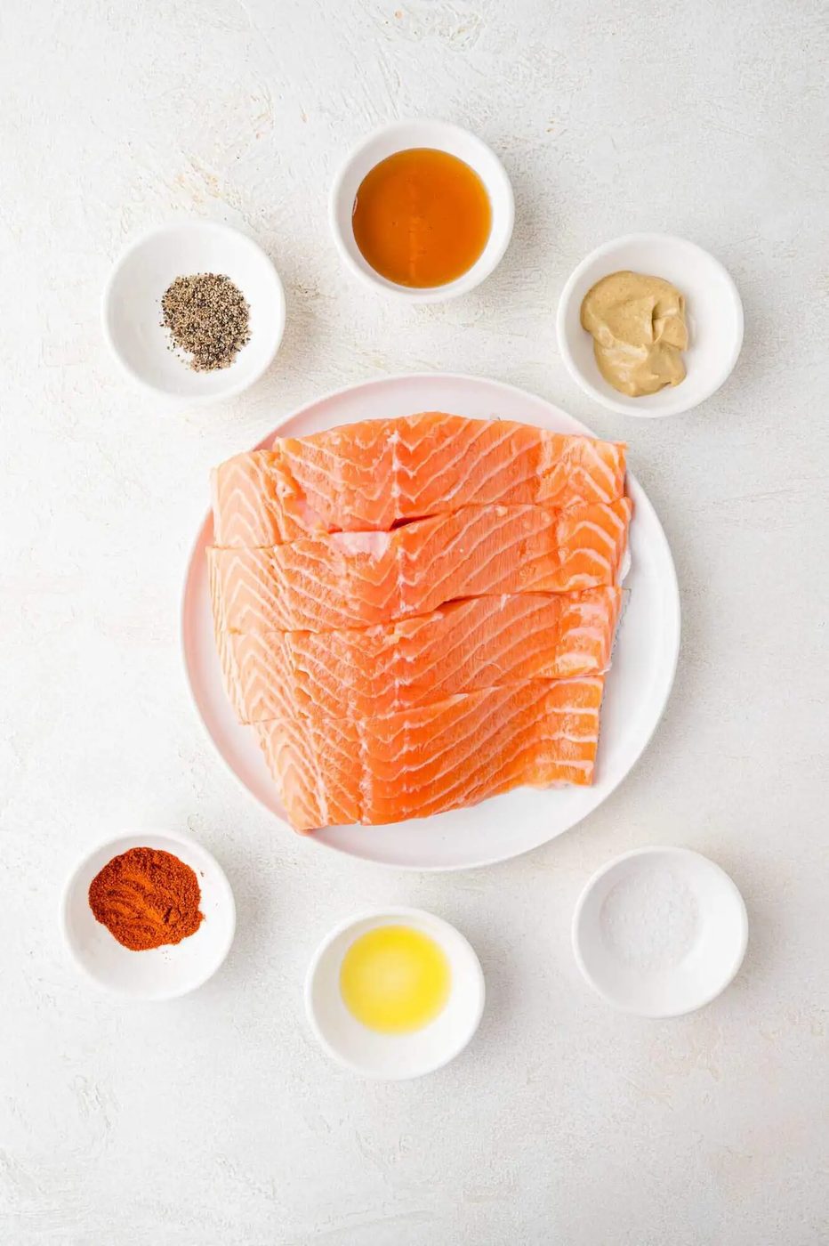 Sliced salmon on a plate surrounded by small bowls containing black pepper, honey, mustard, paprika, and salt.