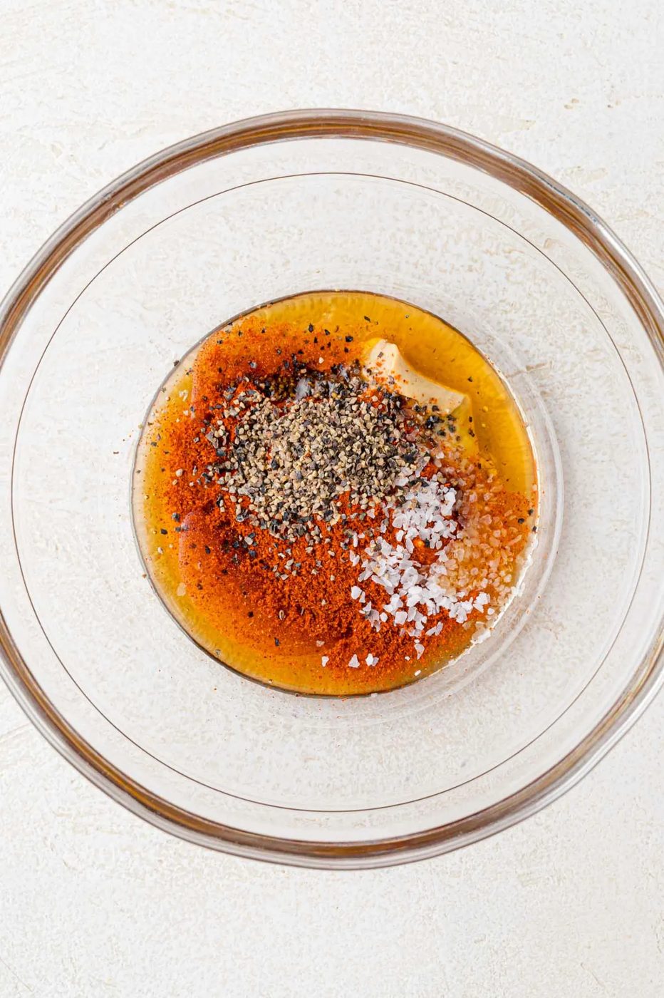 A clear glass bowl containing spices and seasonings, including black pepper, salt, and mustard, on a light textured surface.