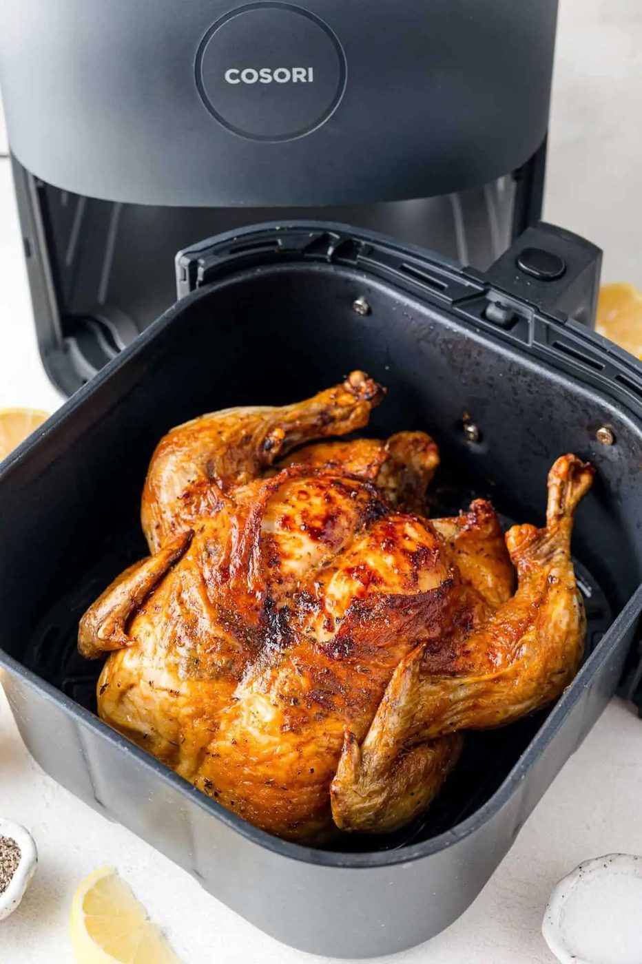 Cooked whole chicken in a black air fryer basket, with the air fryer visible in the background. Lemon slices and seasoning are nearby.