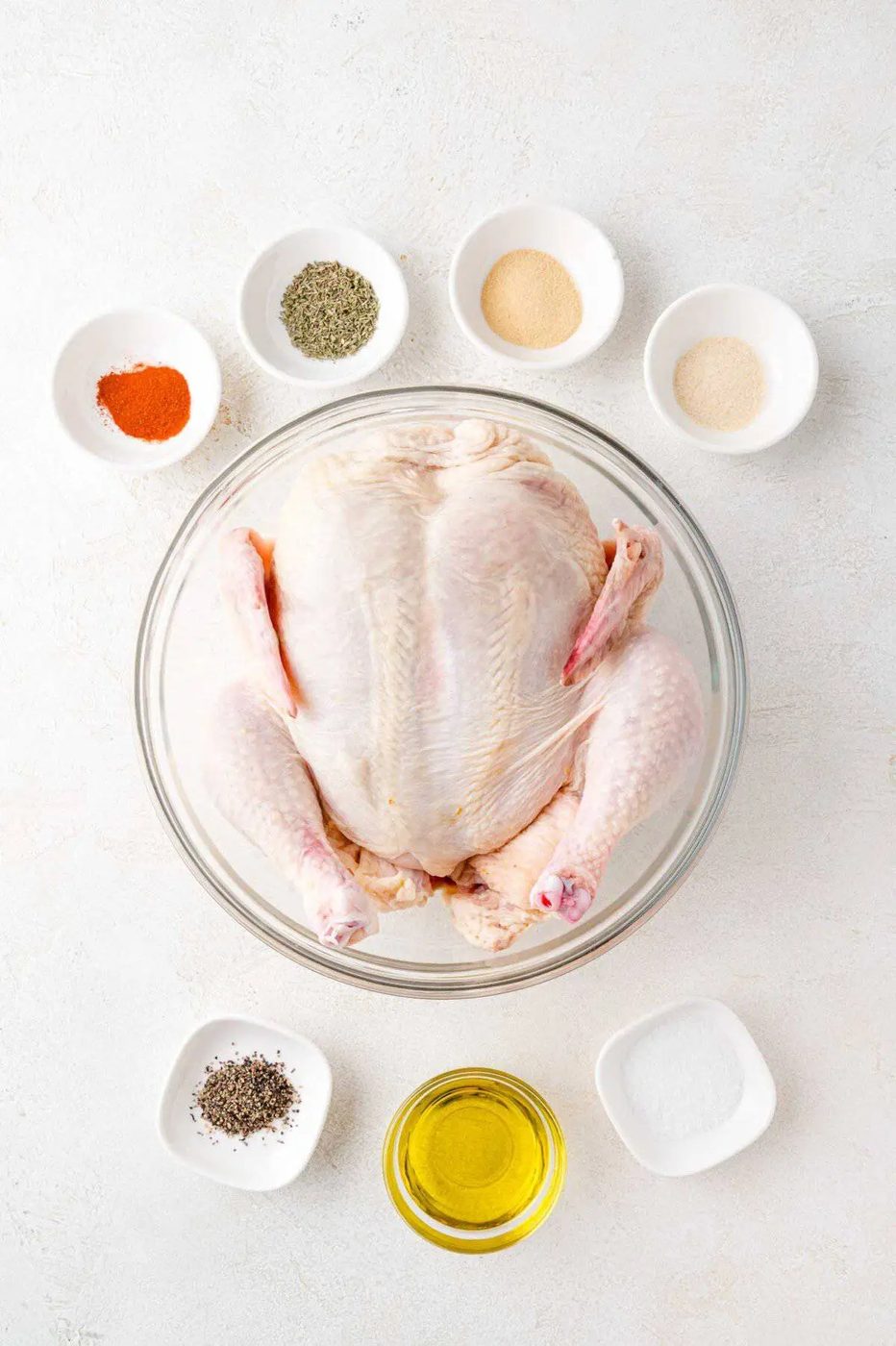 A raw whole chicken in a glass bowl surrounded by small bowls of spices, including black pepper, salt, garlic powder, and oil, on a white surface.