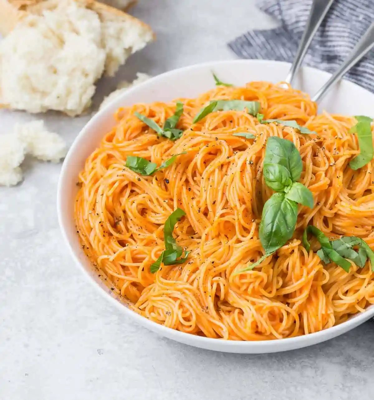 Simple pasta dish made with roasted red peppers and angel hair pasta, in a white bowl, with bread in the background.
