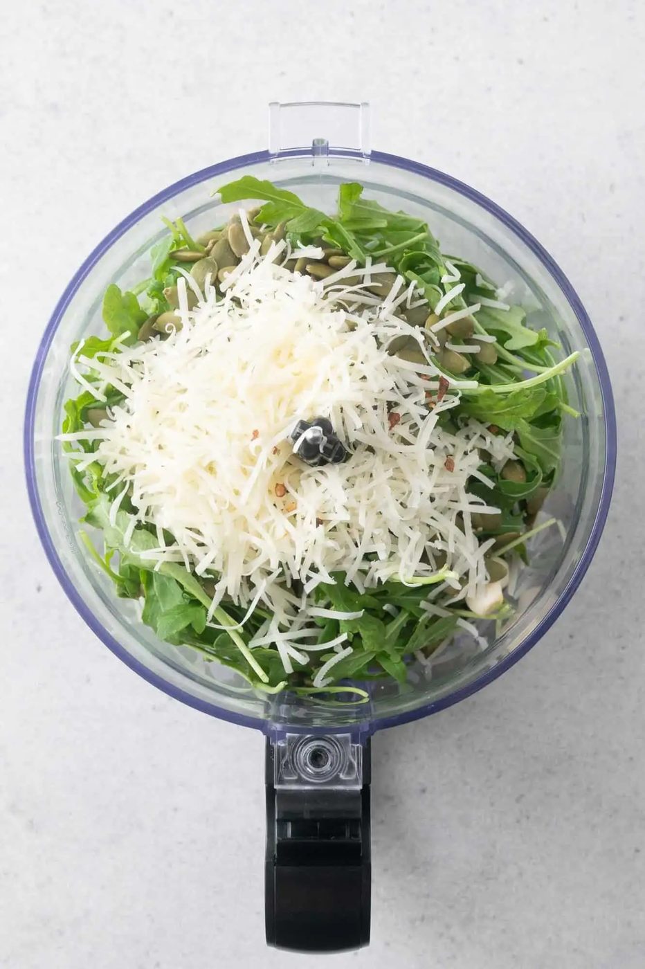 A food processor bowl filled with arugula leaves, grated cheese, pepitas, and seasonings, viewed from above.