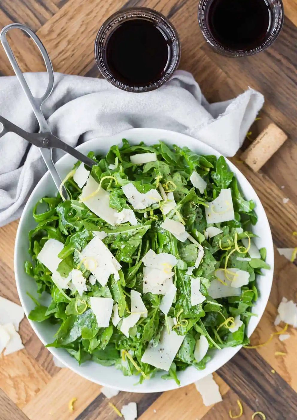 Large bowl of a classic arugula salad with parmesan. Silver serving utensils, red wine, and a cork also pictured.