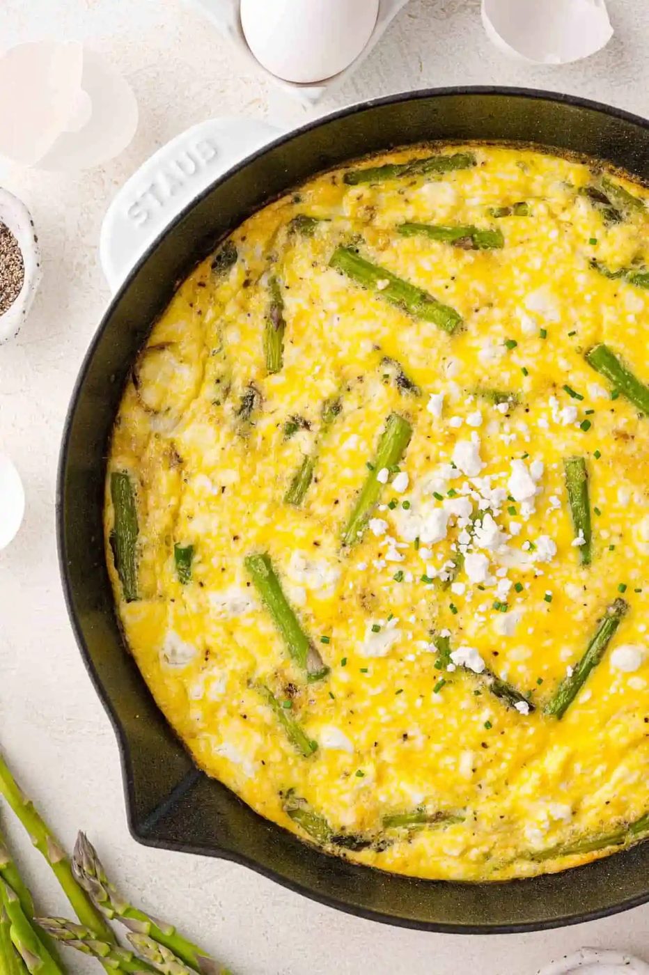 A frittata with asparagus and crumbled goat cheese in a black skillet, surrounded by eggshells and fresh asparagus.