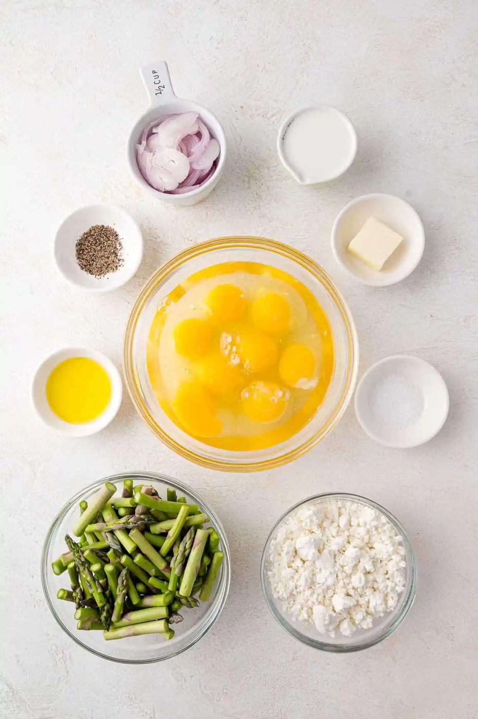 Ingredients on a countertop include eggs, chopped asparagus, goat cheese, shallots, milk, butter, olive oil, salt, and pepper.