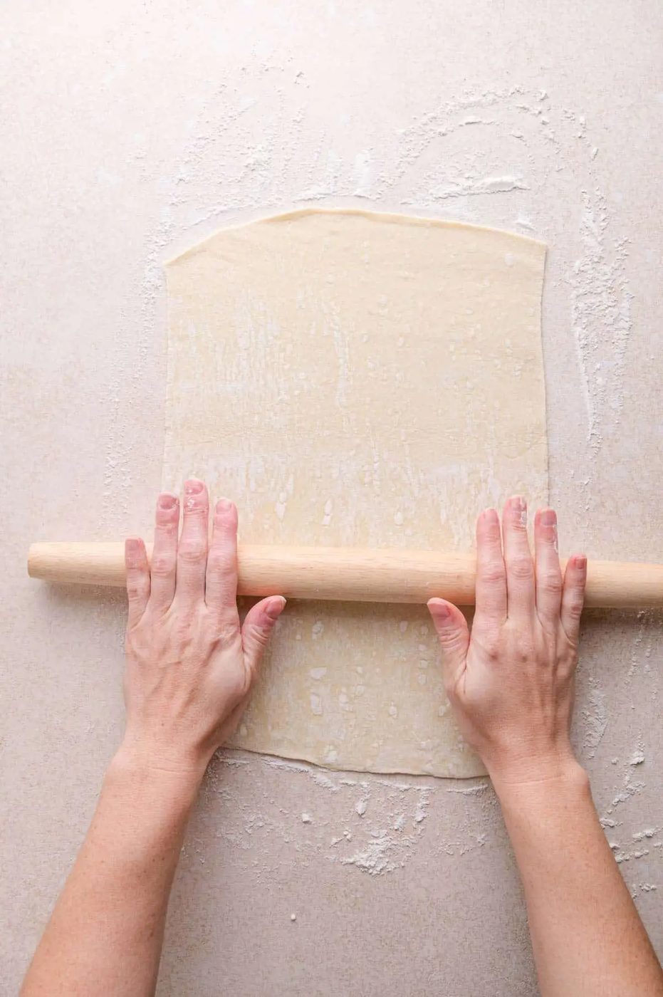 Hands using a rolling pin to flatten puff pastry on a lightly floured surface.