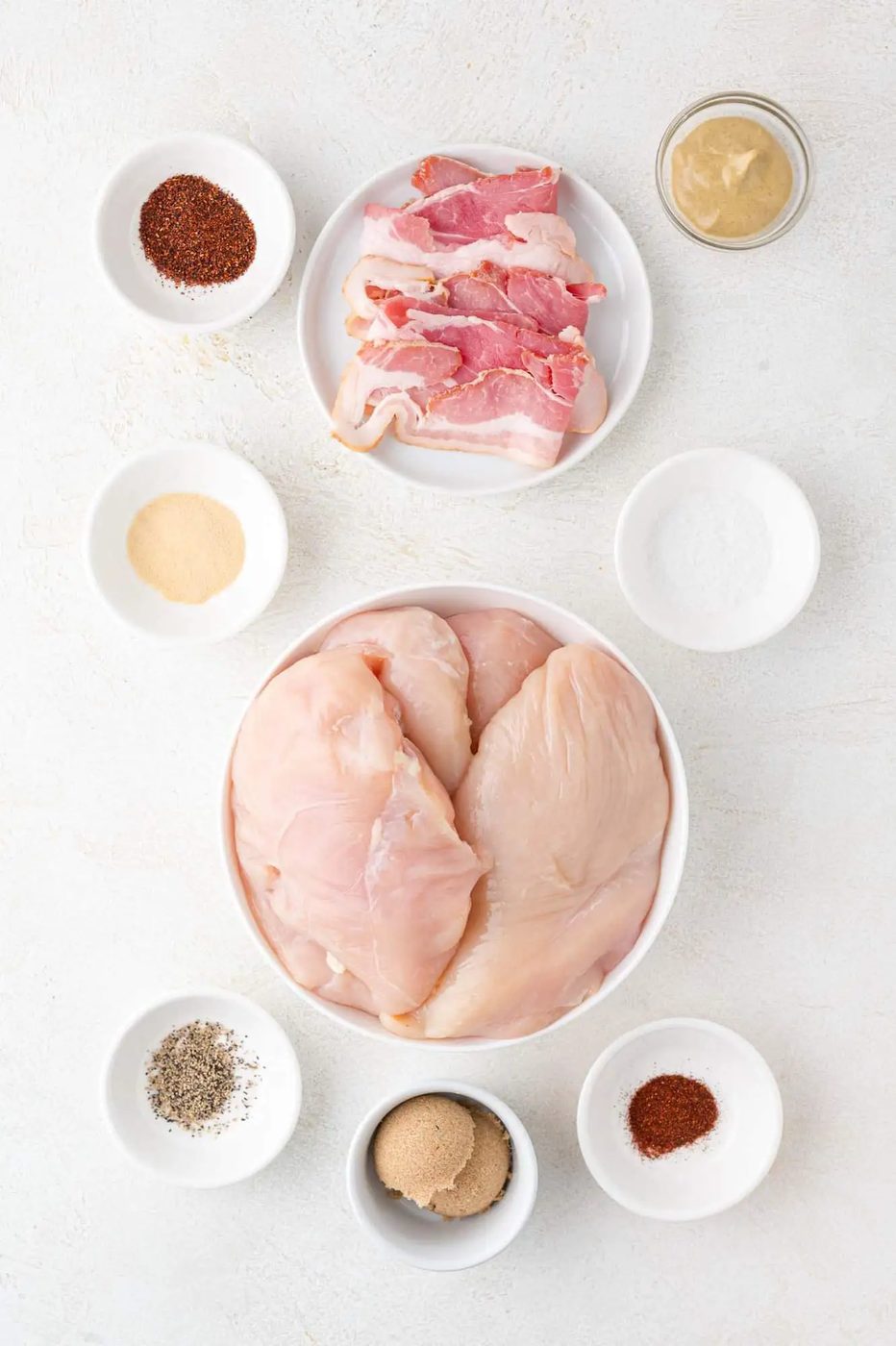 Raw chicken breasts in a bowl surrounded by bowls of bacon, mustard, salt, pepper, brown sugar, garlic powder, paprika, and chili powder on a white surface.