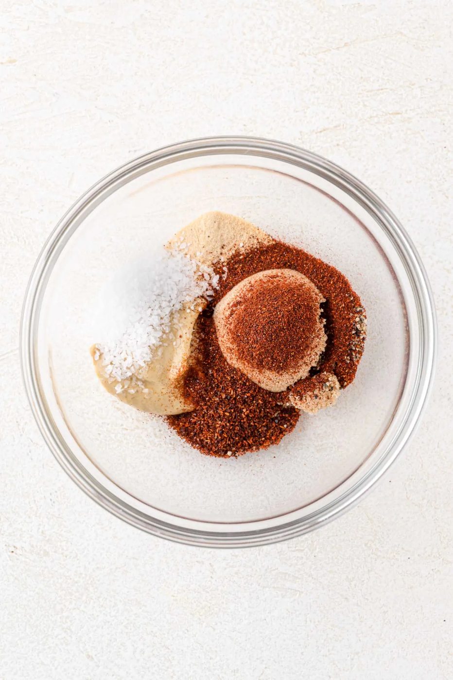A clear glass bowl containing brown sugar, chili powder, salt, and other spices on a white surface.