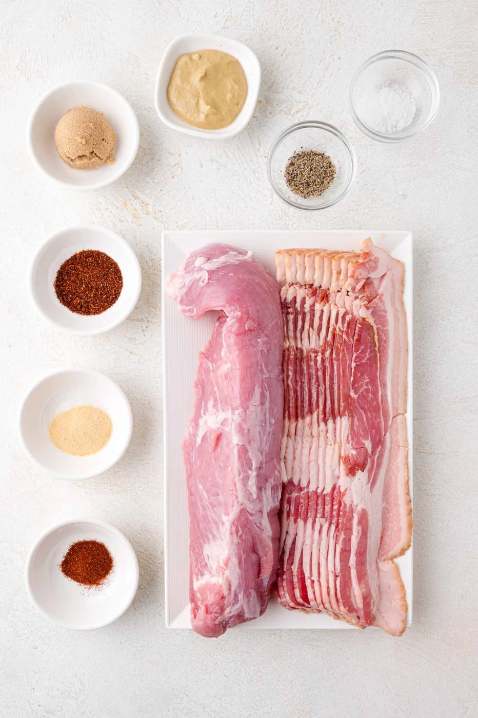 Raw pork tenderloin and bacon on a white tray, surrounded by small bowls of spices and seasonings on a light surface.
