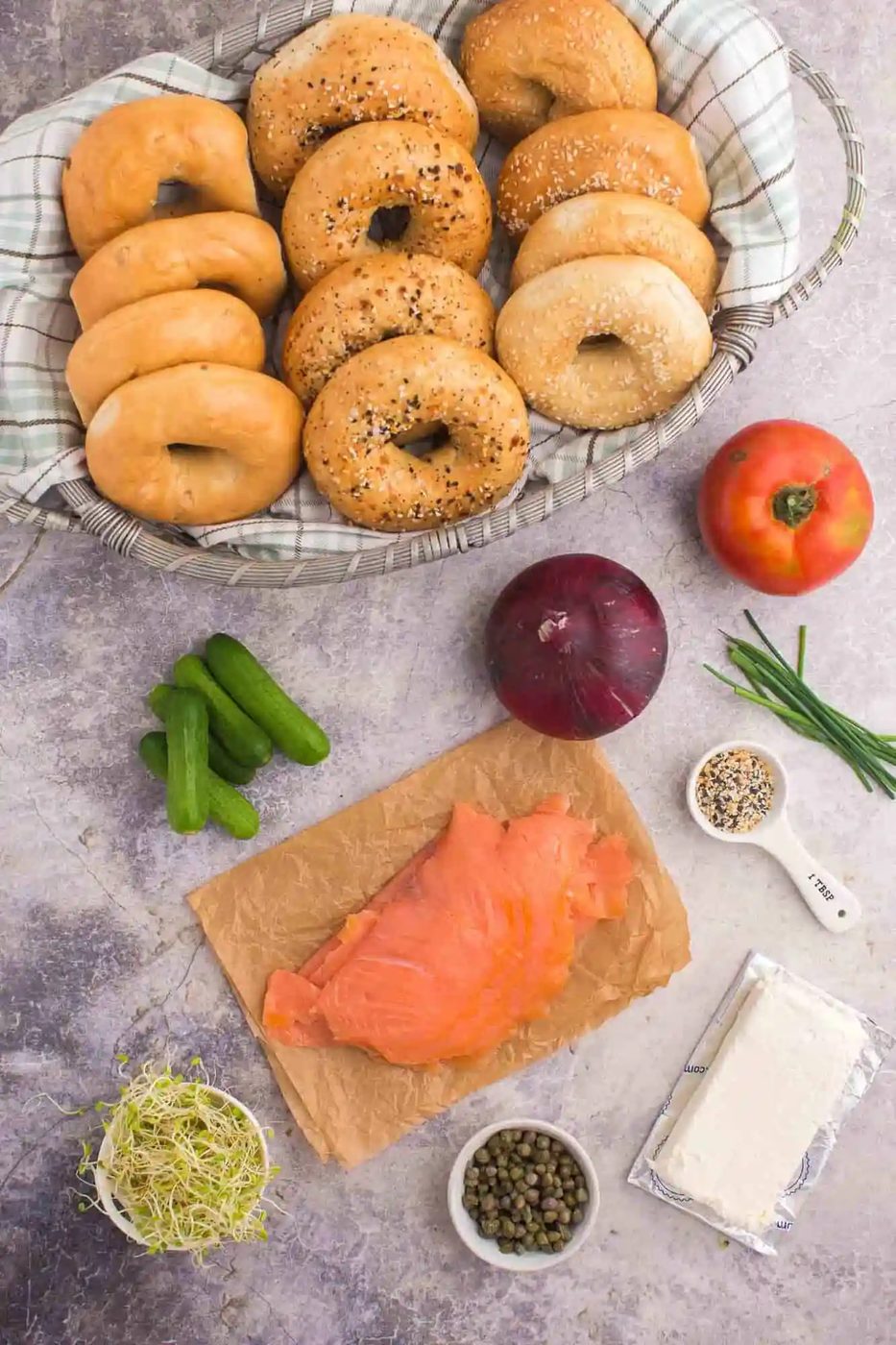Bagels in a basket with smoked salmon, cream cheese, capers, sprouts, red onion, tomato, cucumbers, chives, and seasoning on a countertop.
