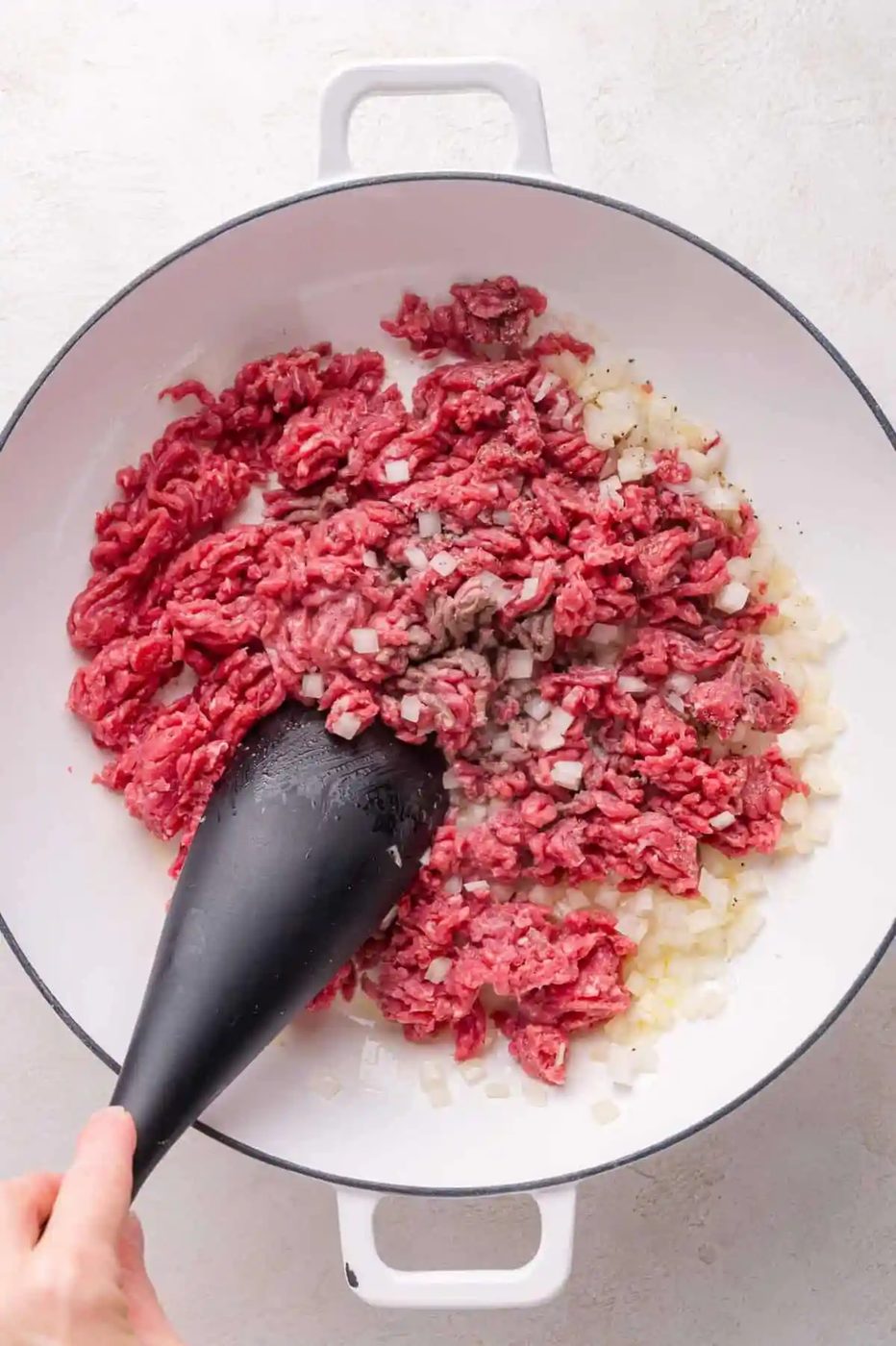 Ground beef and chopped onions being sautéed in a white pan, stirred with a black spatula.