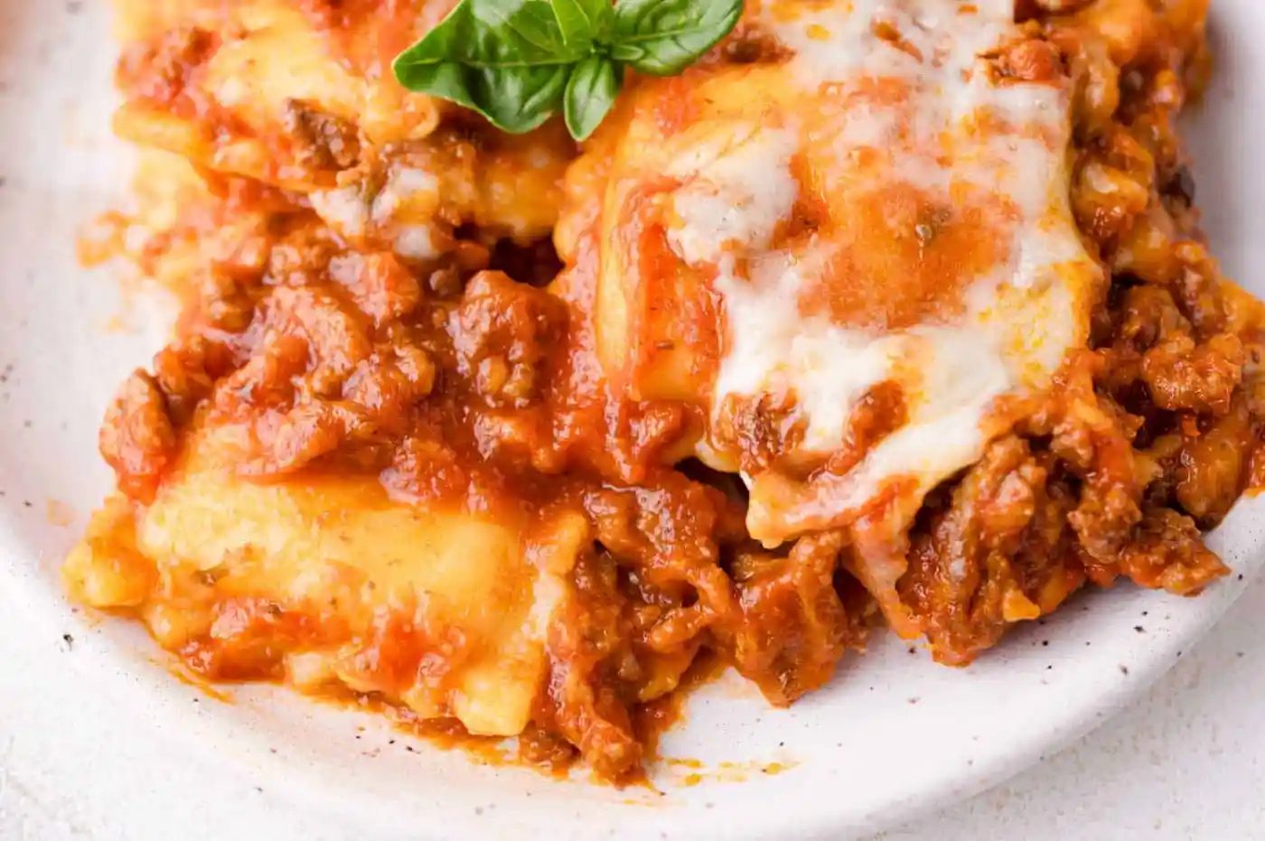 Close-up of a serving of baked ravioli topped with melted cheese and tomato meat sauce, garnished with a fresh basil leaf on a white plate.
