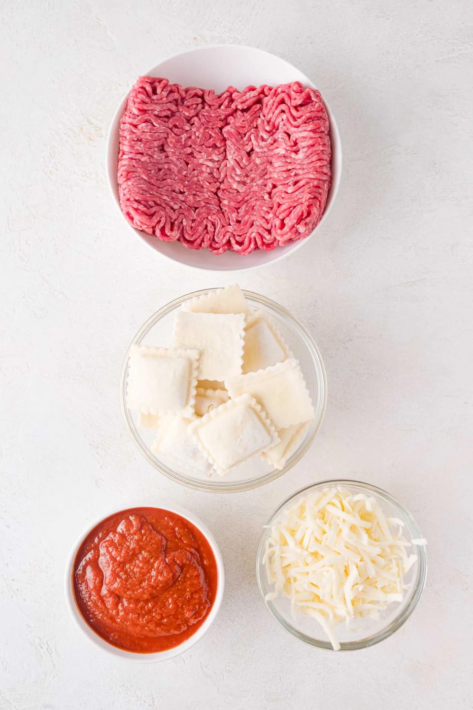 Four bowls contain ground beef, square ravioli, tomato sauce, and shredded cheese, arranged on a white surface.