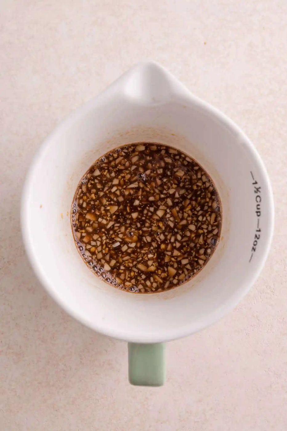 A white measuring cup containing a mixture of finely chopped garlic and balsamic-based sauce, viewed from above on a light surface.