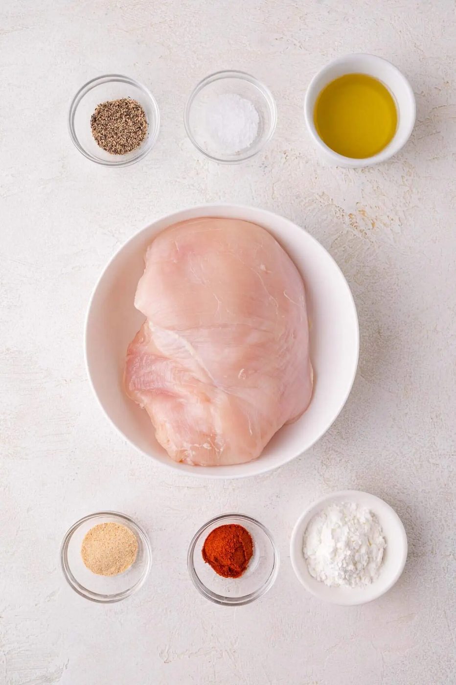 Raw chicken breast in a white bowl surrounded by small bowls containing black pepper, salt, olive oil, garlic powder, paprika, and cornstarch on a light surface.