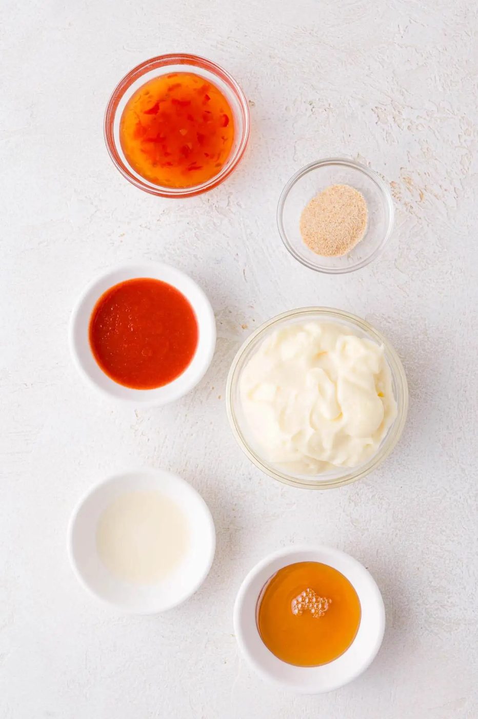 Six small bowls containing mayonnaise, sweet chili sauce, sriracha, garlic powder, vinegar, and honey are arranged on a white surface.