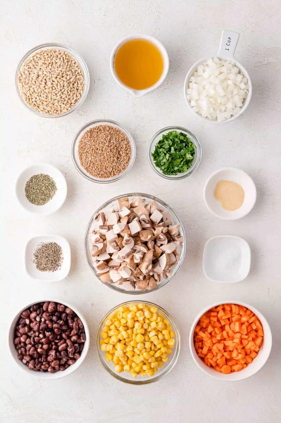 Ingredients arranged on a white surface: barley, broth, diced onions, mushrooms, black beans, corn, carrots, spices, and herbs in bowls.