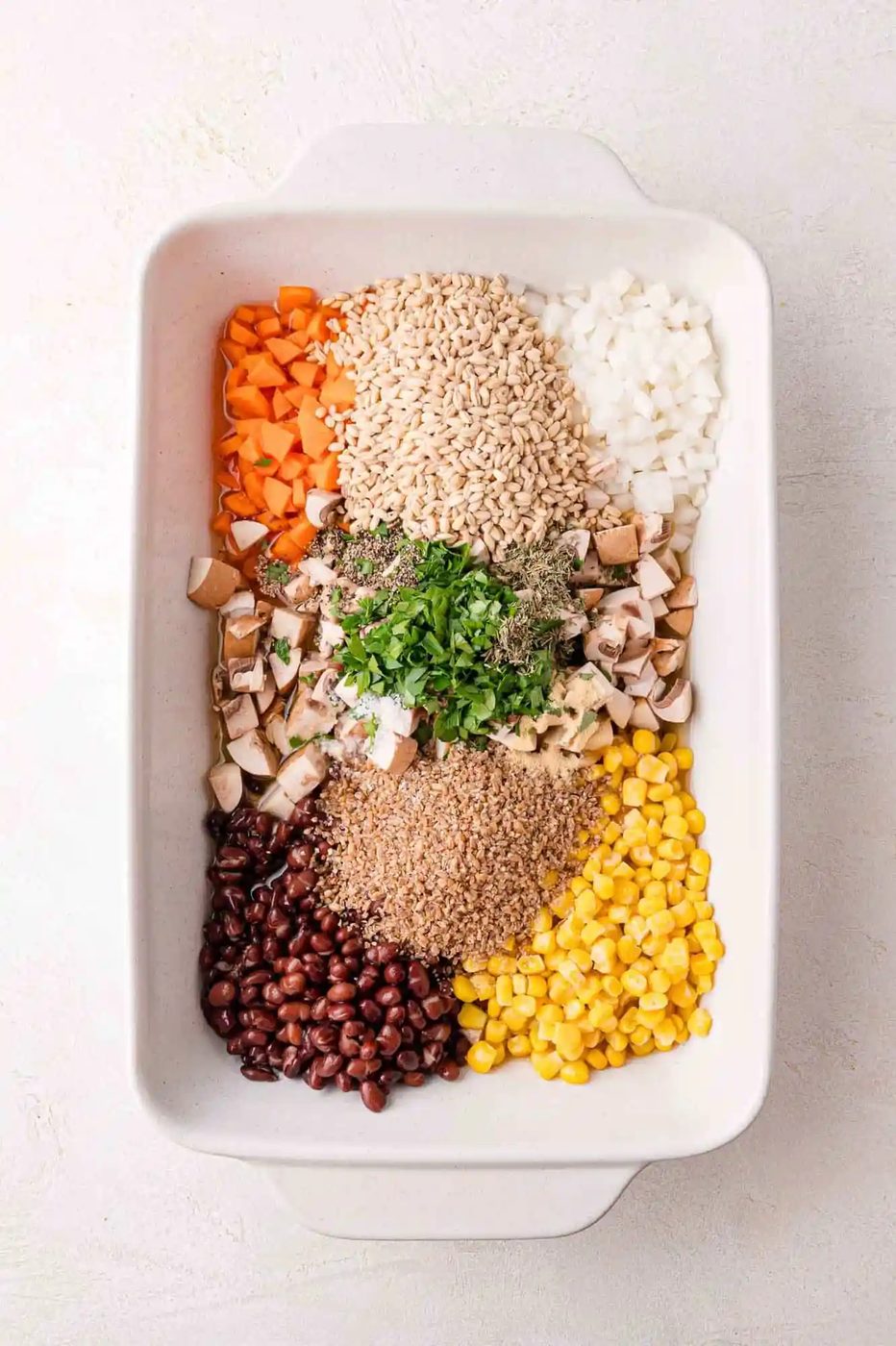 A white dish with uncooked ingredients: barley, diced onion, diced carrot, black beans, corn, chopped mushrooms, herbs, and greens arranged in separate sections.