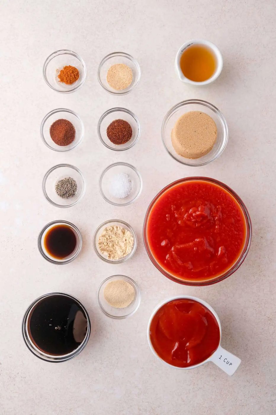 Ingredients for from-scratch barbecue sauce arranged on a white surface, including spices, brown sugar, vinegar, Worcestershire sauce, molasses, tomato sauce, and crushed tomatoes.
