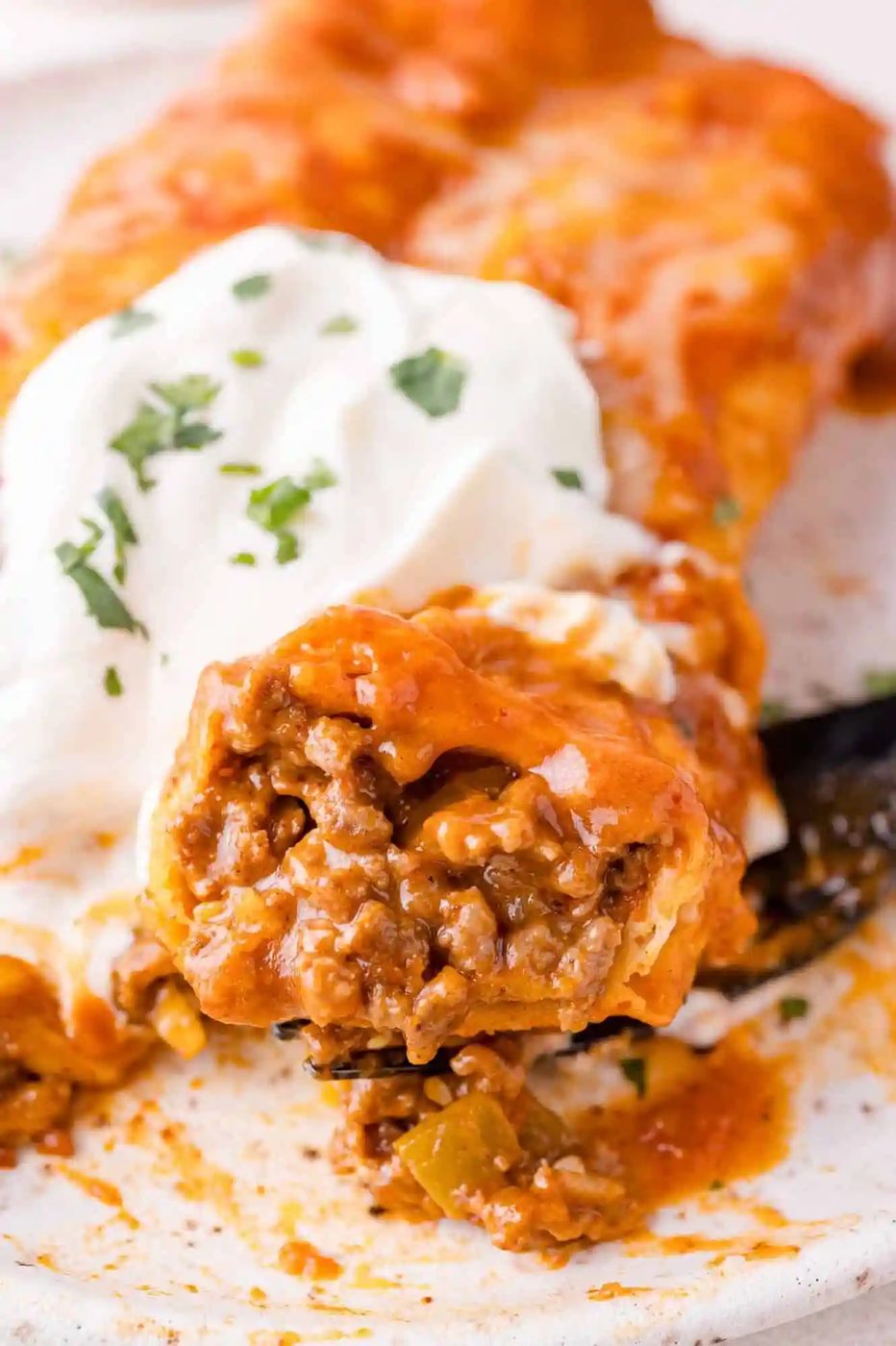 Close-up of a beef enchilada topped with melted cheese and sour cream, garnished with chopped herbs.
