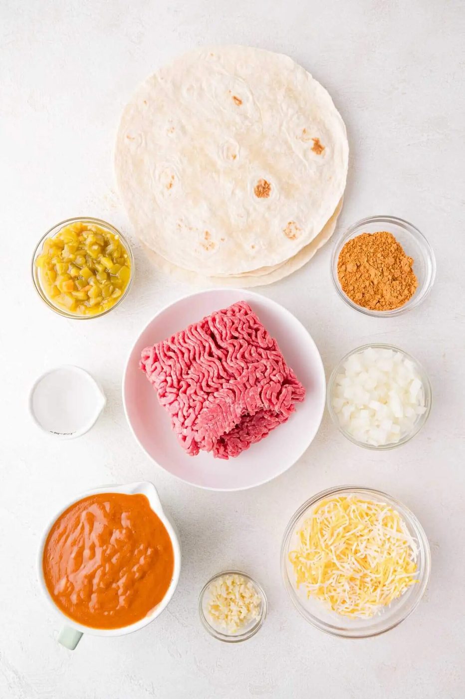 Ingredients for enchiladas include ground beef, tortillas, cheese, chopped onion, sauce, sour cream, refried beans, garlic, taco seasoning, and green chilies on a white surface.