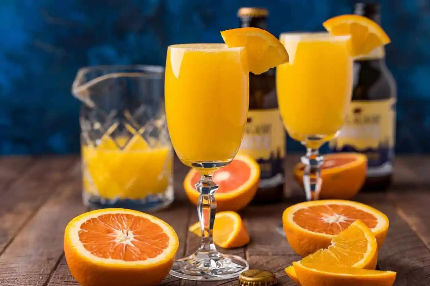 Two glasses of orange beermosa garnished with orange slices sit on a wooden table with halved oranges, a pitcher of juice, and bottles in the background.