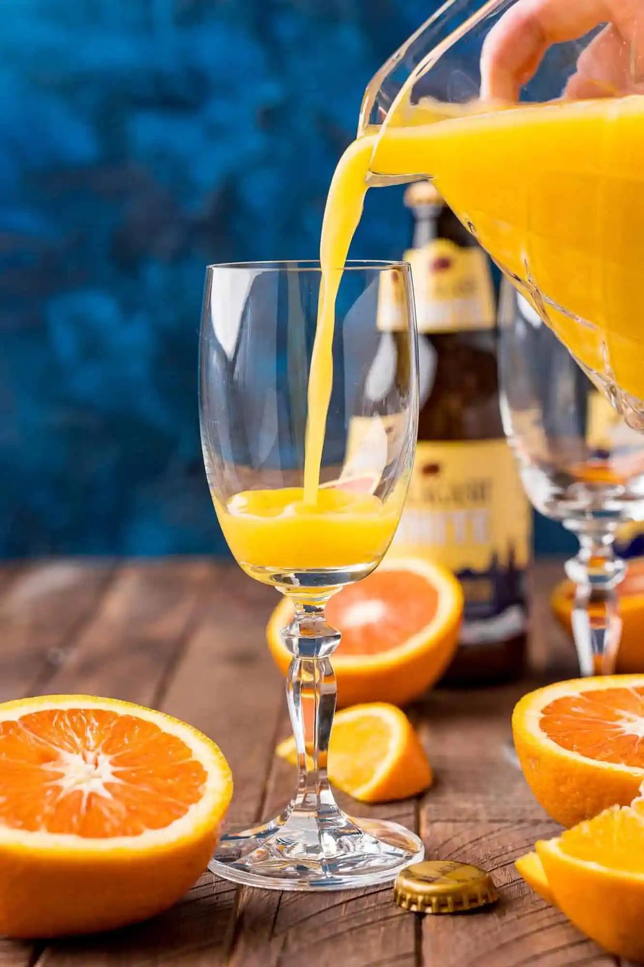 A hand pours orange juice from a pitcher into a wine glass on a wooden table with sliced oranges and a beer bottle in the background.