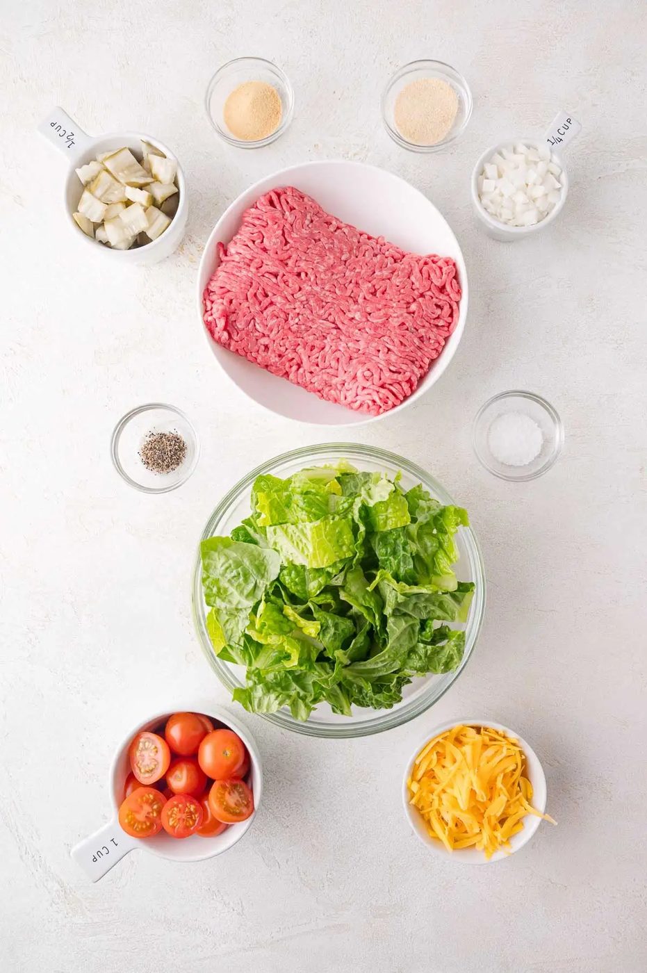 Ingredients on a table including ground beef, lettuce, cherry tomatoes, cheddar cheese, diced onions, seasonings, and dill pickles, ready for preparation.
