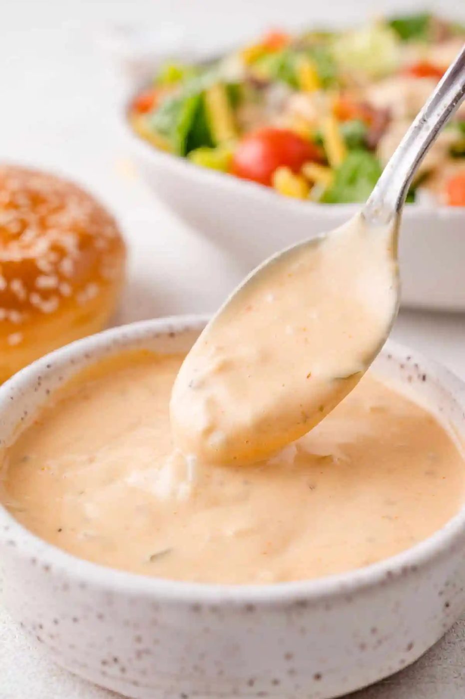 Spoon dipping into a bowl of creamy pink sauce, with a salad and a burger bun in the background.