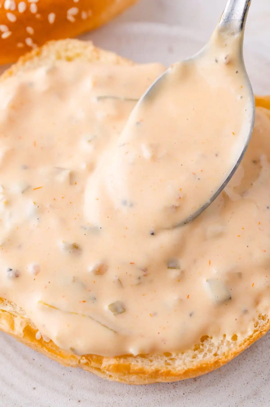 A spoon spreading creamy sauce with chopped pickles on a halved sesame seed burger bun placed on a white plate.