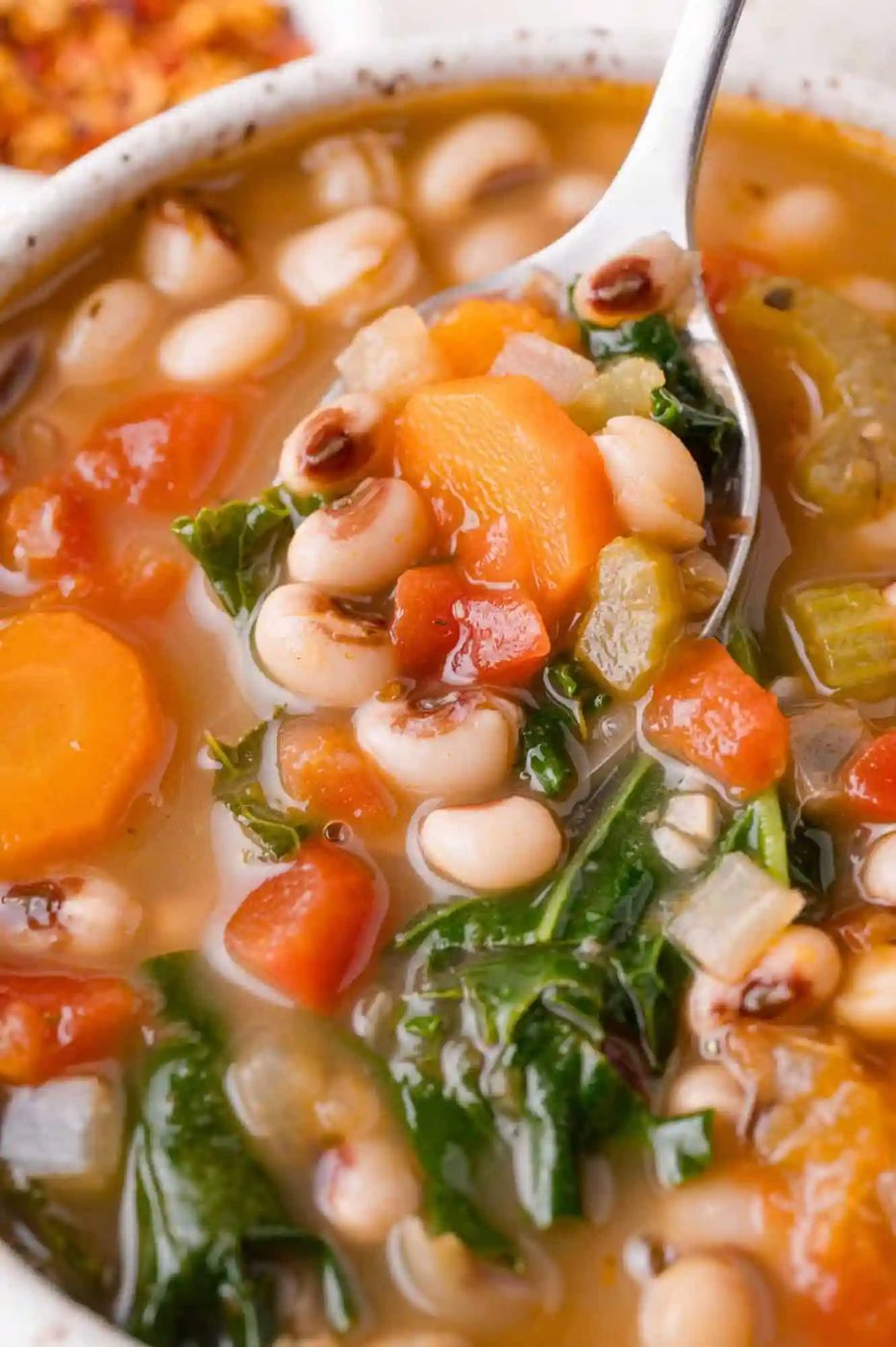 A close-up of a bowl of black-eyed pea soup with carrots, tomatoes, celery, and leafy greens, with a spoon lifting a portion.