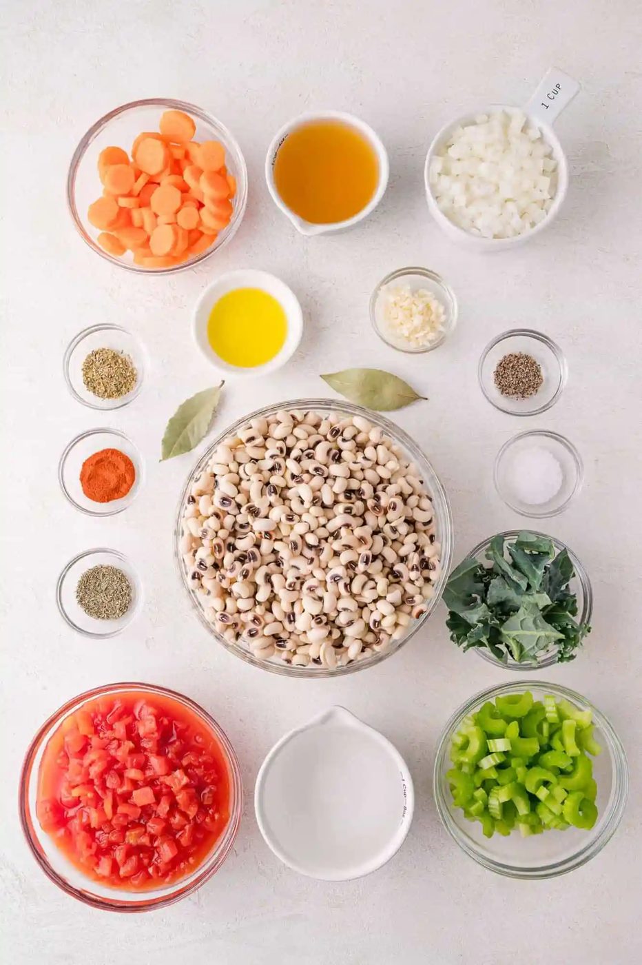 Overhead view of prepped ingredients in bowls, including black-eyed peas, diced tomatoes, carrots, celery, onion, garlic, kale, spices, bay leaves, broth, and oil on a white surface.