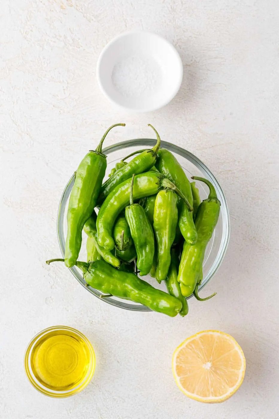 A bowl of green shishito peppers on a white surface, with a halved lemon, a small bowl of olive oil, and a small bowl of salt nearby.