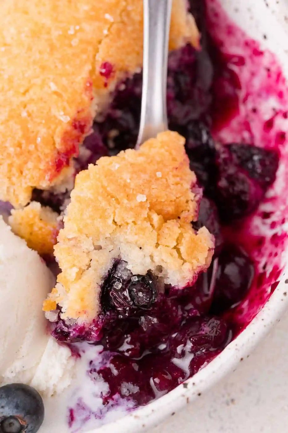 A close-up of a blueberry cobbler with a golden biscuit topping, a spoonful lifted, served with a scoop of vanilla ice cream on the side.