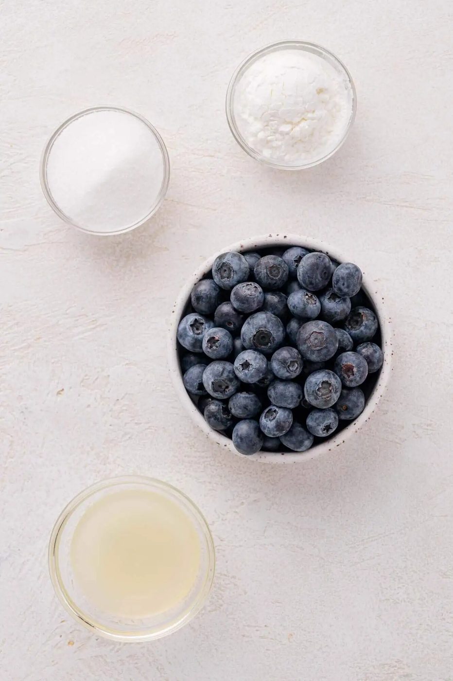 A bowl of blueberries, a small bowl of sugar, a small bowl of cornstarch, and a small cup of lemon juice on a light surface.