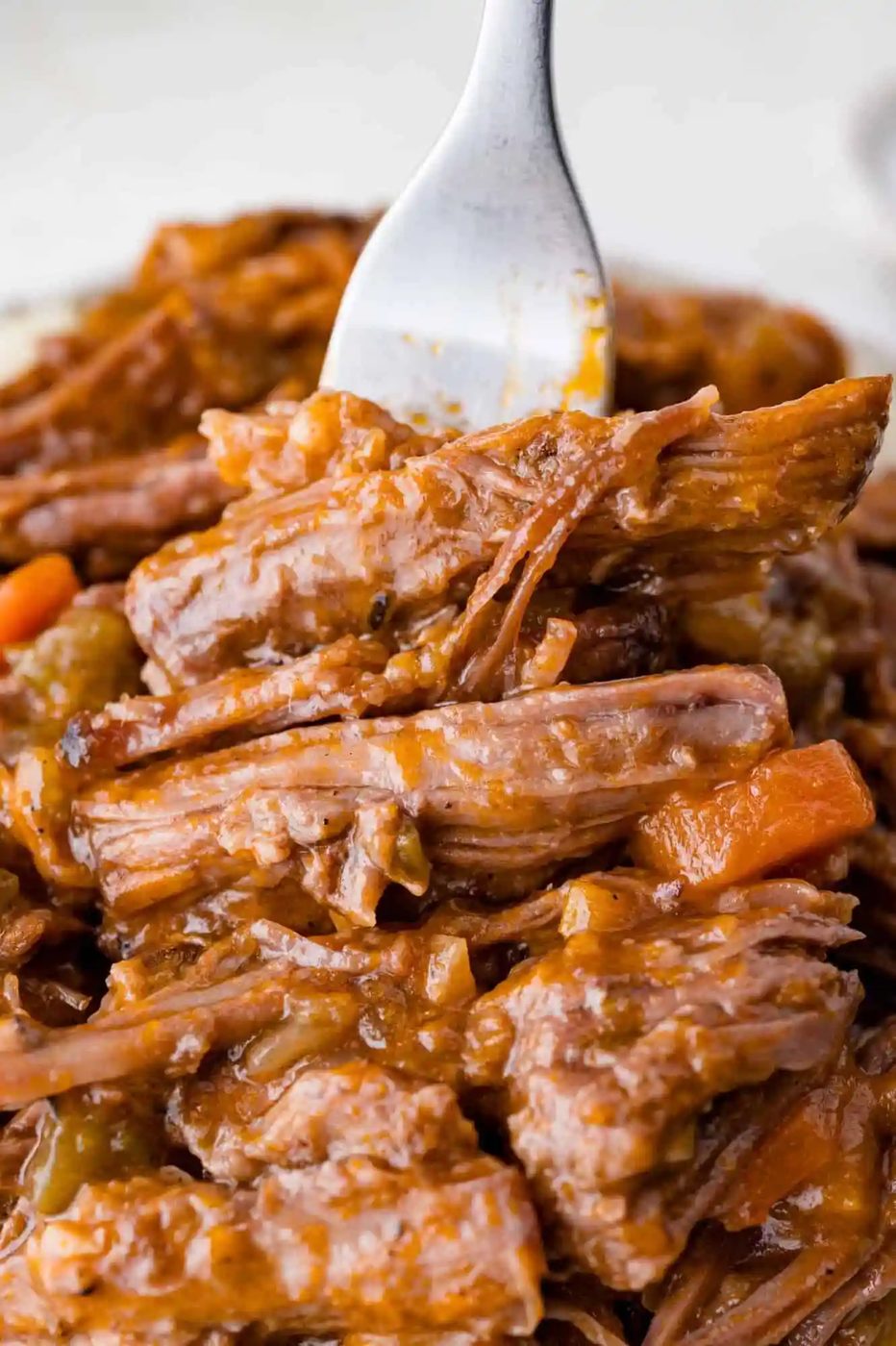 A fork holds a portion of shredded braised beef covered in gravy, with visible pieces of carrot and onion.