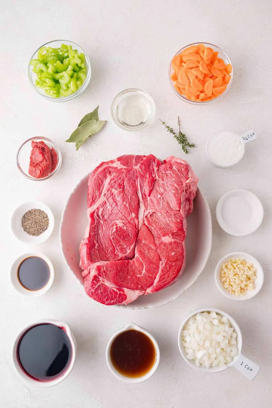 Raw beef on a plate surrounded by ingredients like chopped celery, carrots, onions, garlic, tomato paste, bay leaf, thyme, red wine, broth, and seasoning in bowls on a white surface.