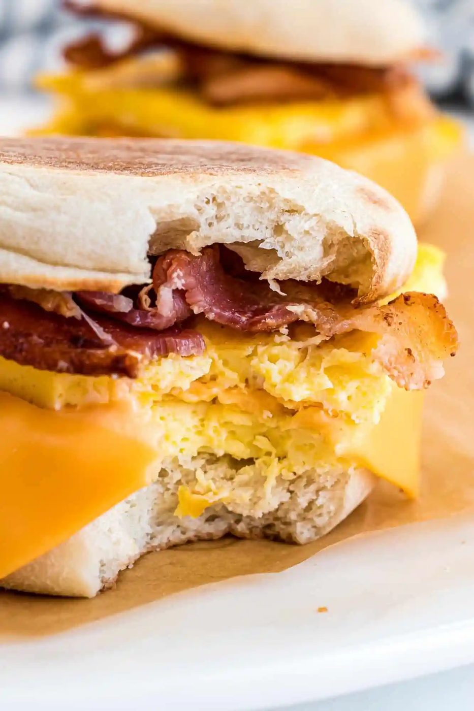 Breakfast sandwich with bacon, scrambled eggs, and melted cheese on an English muffin. A bite is taken out of one sandwich.