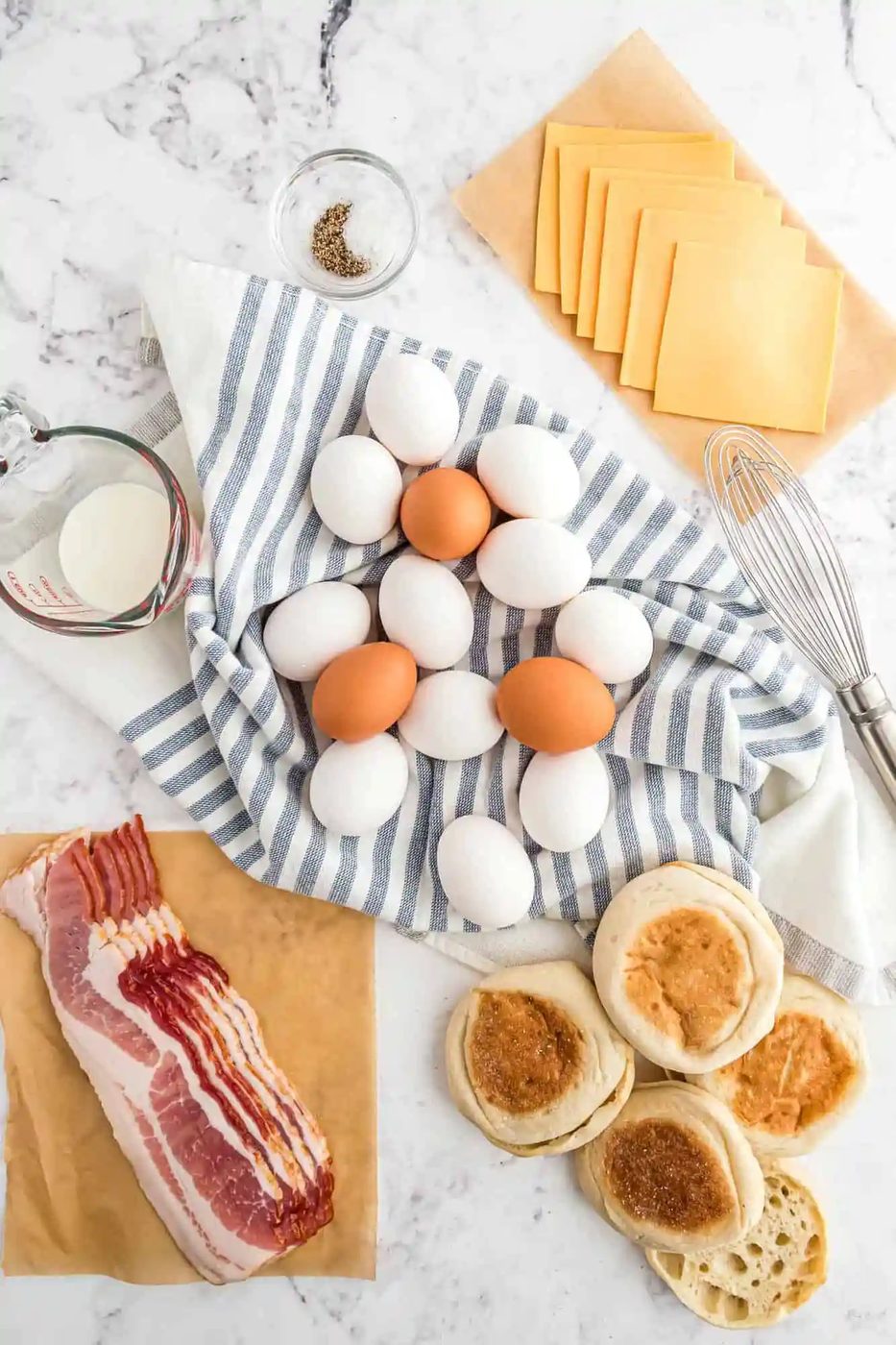 Eggs on a striped cloth, bacon, cheese slices, English muffins, a whisk, a cup of milk, and a small bowl of salt and pepper on a marble surface.