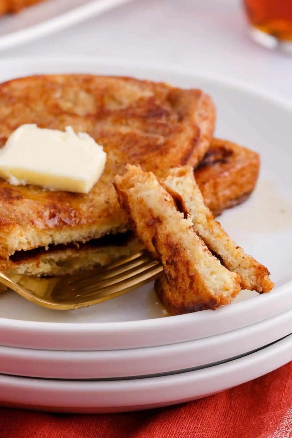A stack of French toast topped with a butter pat sits on a white plate. A fork holds a cut piece of toast in front.