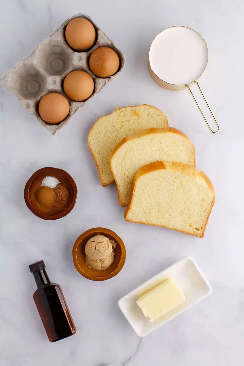 Ingredients for French toast: eggs in a carton, slices of bread, a cup of milk, butter, vanilla extract, brown sugar, and salt in bowls on a marble surface.