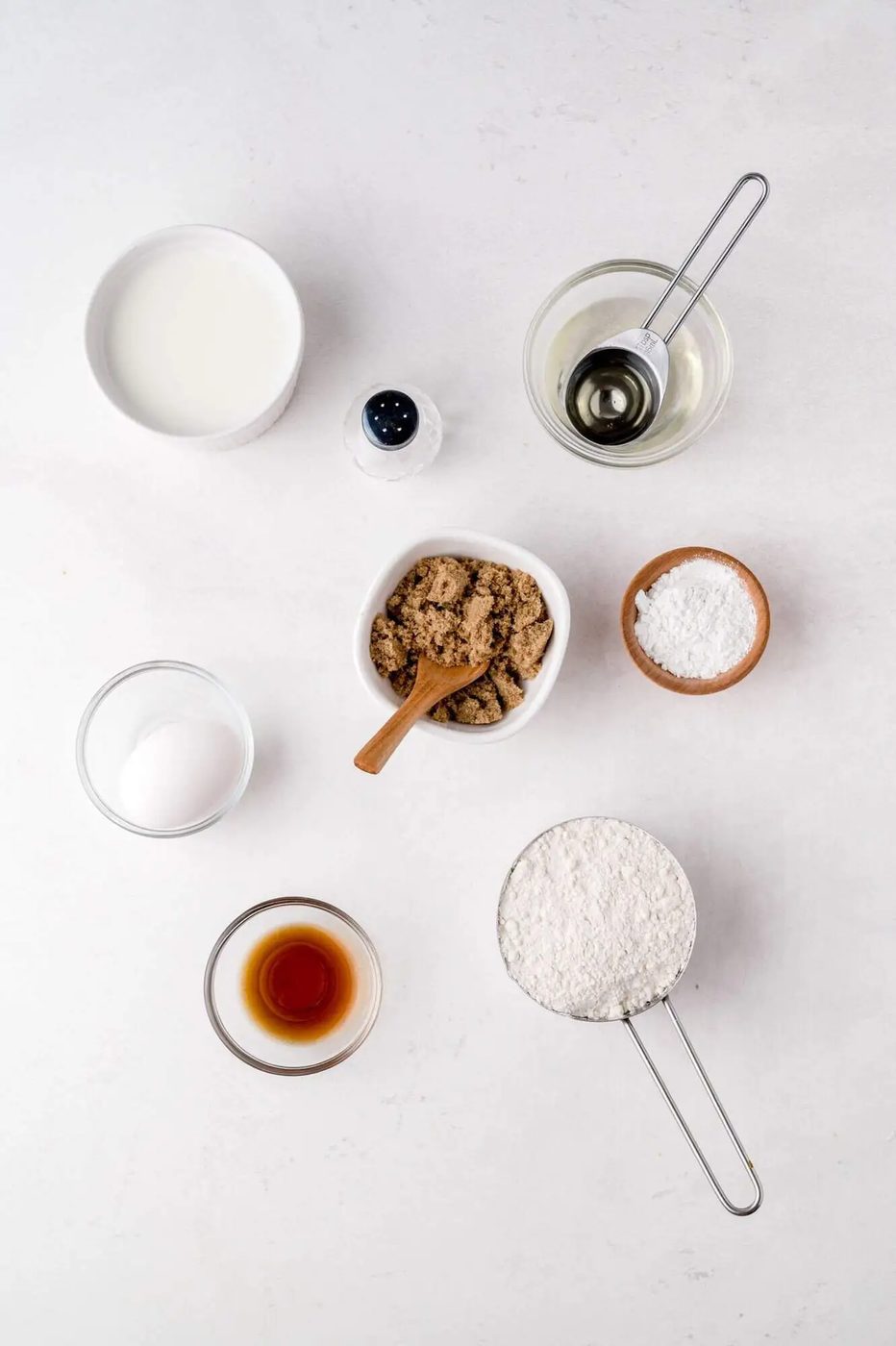 Ingredients arranged on a white surface, including milk, an egg, brown sugar, vanilla extract, flour, oil, salt, and baking soda in various bowls and measuring tools.