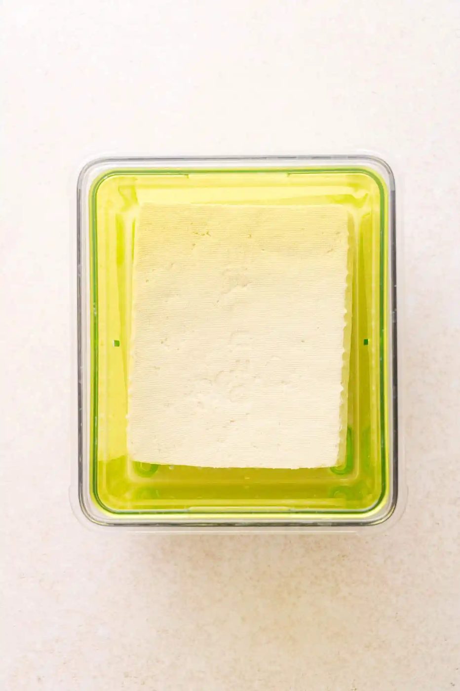 A block of tofu sits in a transparent container with a green base, filled with liquid, viewed from above on a light surface.