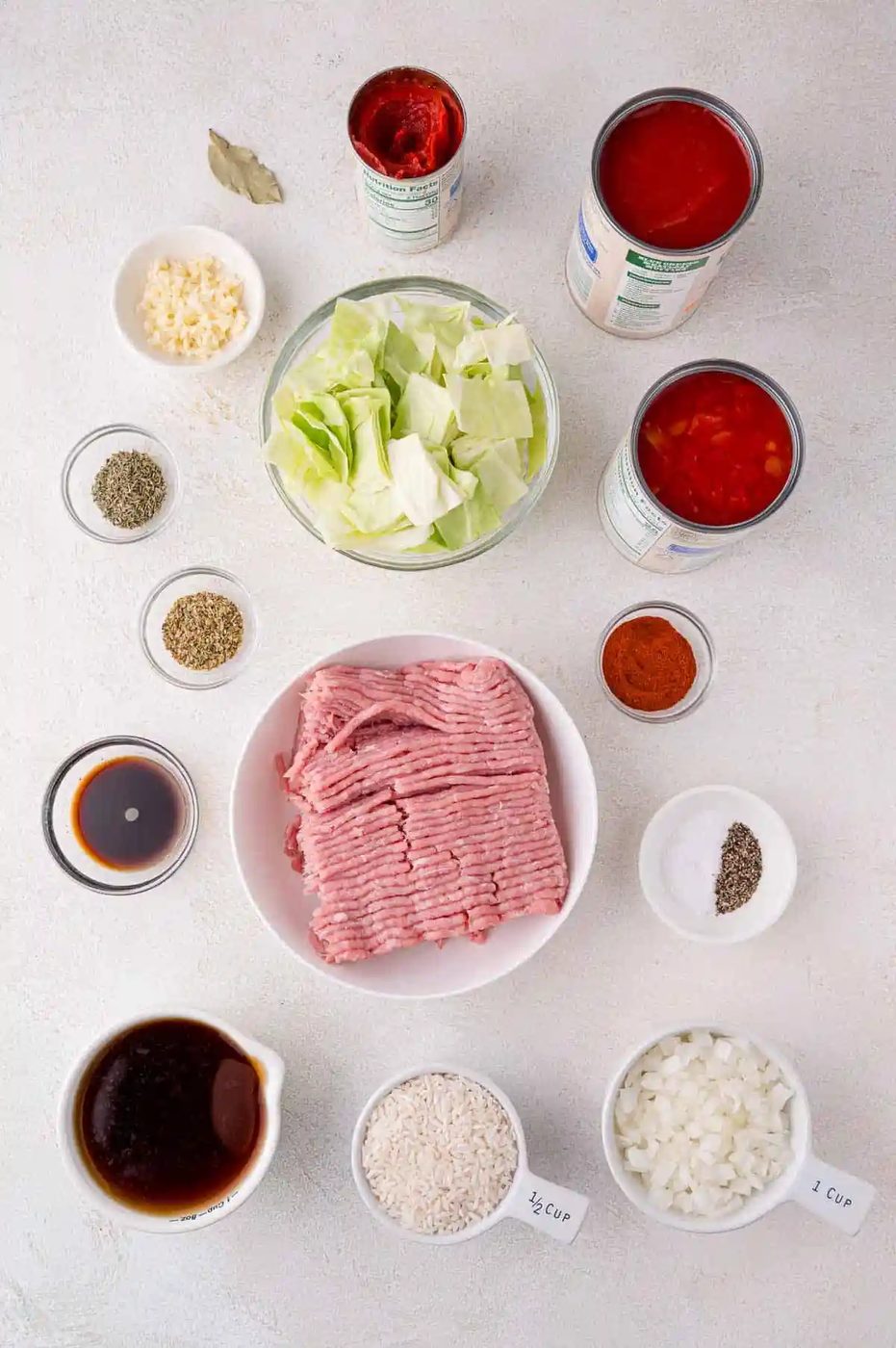 Ingredients for cabbage roll soup, including ground meat, chopped cabbage, canned tomatoes, rice, onions, garlic, and various seasonings, arranged on a light surface.