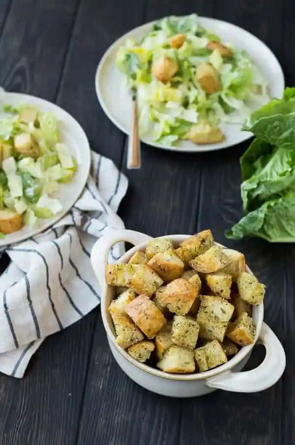 Bowl with Caesar croutons in foreground, two Caesar salads in background.