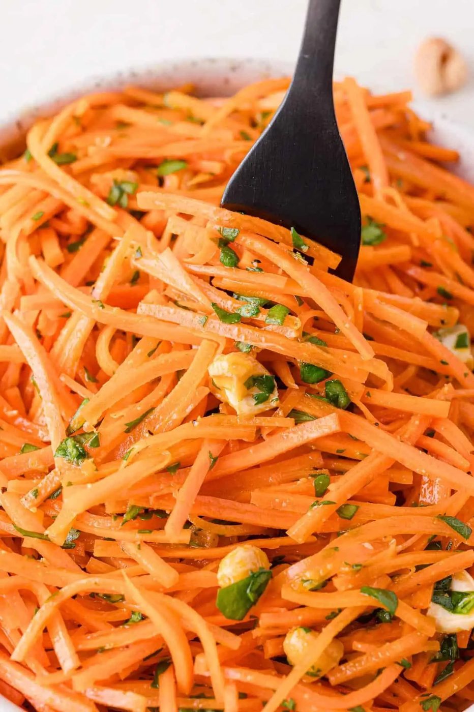 A close-up of a carrot salad with chopped herbs and nuts, with a black fork in the dish.