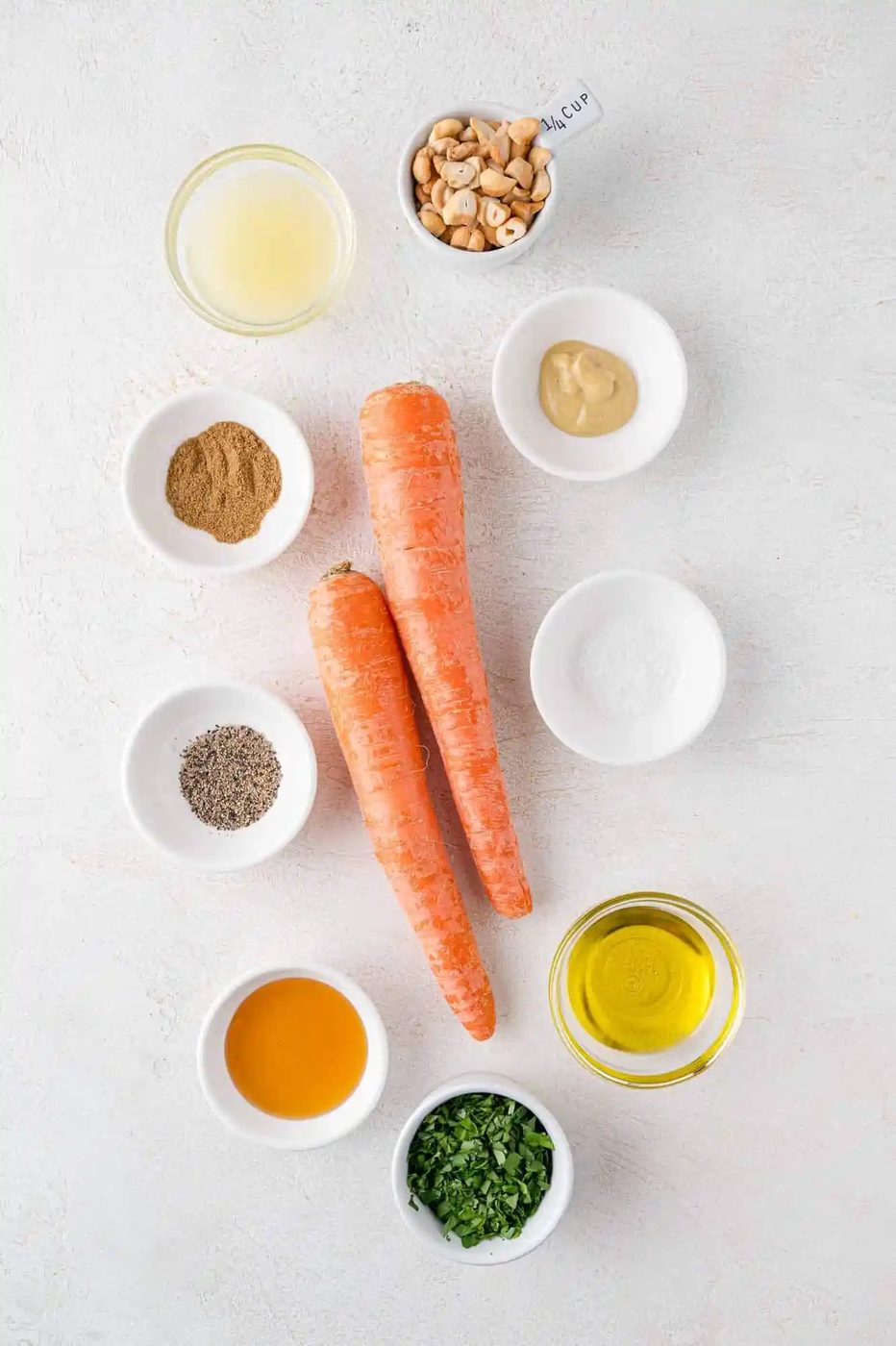 Ingredients flat lay: two carrots, bowls of mustard, olive oil, lemon juice, cilantro, sugar, cashews, ground spice, salt, pepper, and honey on a light background.