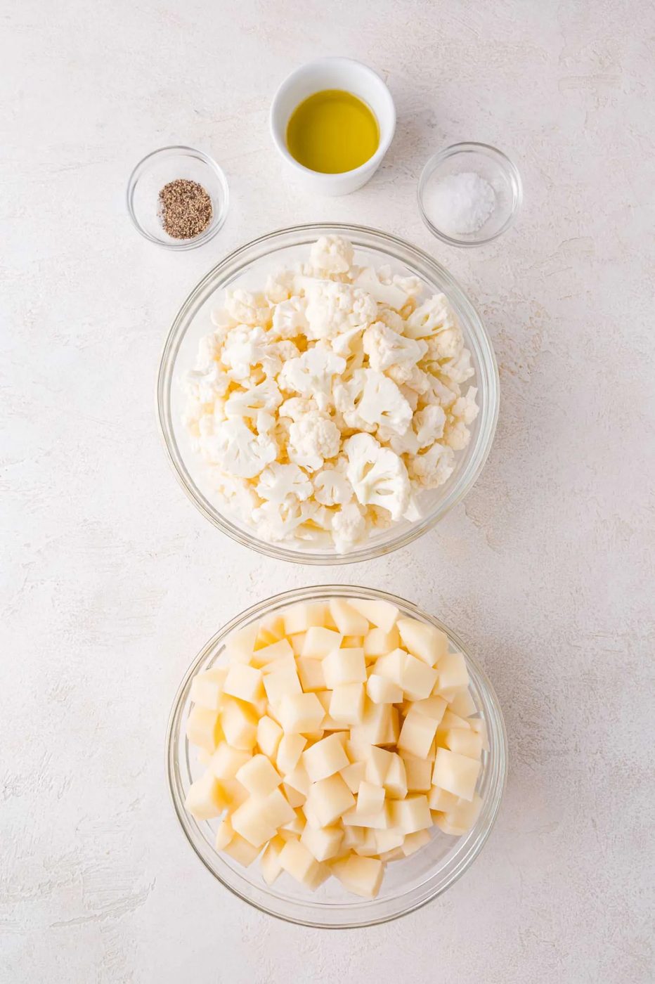 Two bowls containing cauliflower florets and diced potatoes, with small bowls of olive oil, salt, and pepper arranged above them on a light surface.