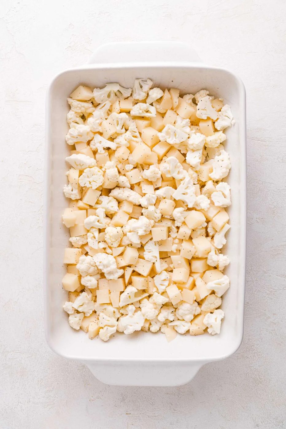 Chopped cauliflower and potatoes in a white rectangular baking dish on a light surface.